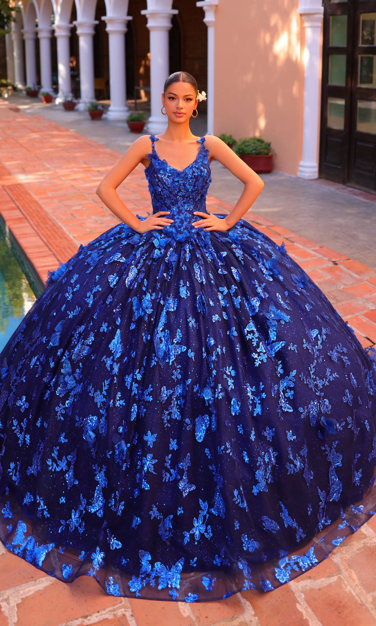 A young woman poses outdoors in front of arched architecture, wearing the Amarra 54260 Corset Quincea¤era Dress with Cloak, featuring a voluminous dark blue design and intricate blue floral patterns.